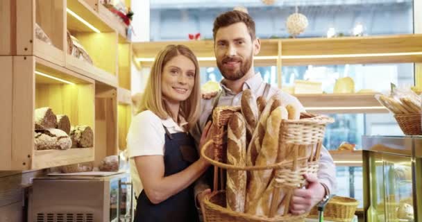 Portret van vrolijke mooie blanke jonge familie getrouwd paar bakkers in goed humeur mand met vers brood staan in kleine eigen bakkerij winkel en glimlachen naar de camera. Begrip familiebedrijf — Stockvideo