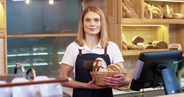 Porträt einer fröhlichen schönen jungen kaukasischen Verkäuferin Bäckereibesitzerin in Schürze, die in einem kleinen Laden steht und einen Korb mit frisch gebackenem Brot in der Hand hält, in die Kamera schaut und allein lächelt — Stockvideo
