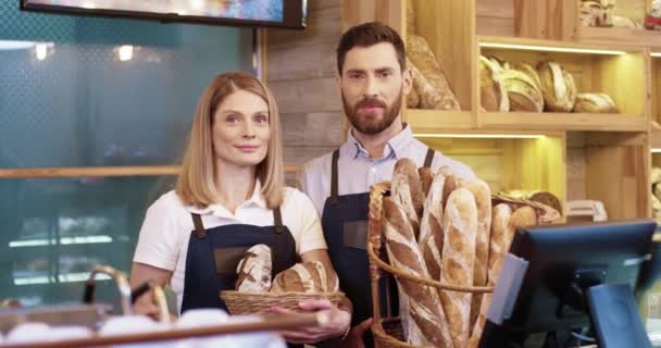Blisko radosnej szczęśliwej młodej kaukaskiej pary małżeńskiej w dobrym nastroju trzymającej kosze ze świeżym chlebem stojącym w małym sklepie piekarniczym i uśmiechniętym do kamery. Mężczyzna i kobieta sprzedawca w piekarni — Wideo stockowe