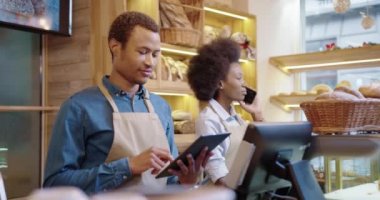 Güzel bir bayan iş arkadaşı cep telefonuyla konuşurken fırında yazı yazan ve tableti tıkırdatan mutlu Afrikalı Amerikalı erkek işçiyi kapatın. Girişimciler konsepti