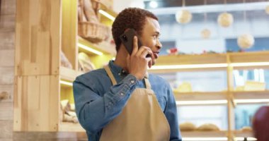Afrika kökenli Amerikalı genç ve mutlu erkek işçinin önlüklü hali fırında duruyor ve akıllı telefondan sohbet ediyor. Keyifli bir şekilde telefon ediyor ve kol saatine bakıyor. Satıcı konsepti. Perakende sektörü