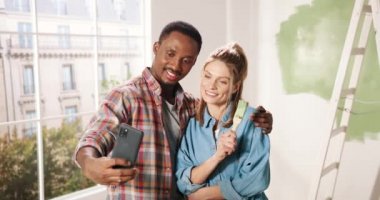 Mutlu melez evli çiftler gülümseyen ve fırçalarla poz veren ve cep telefonuyla fotoğraf çeken genç çiftler. Ev onarımı konsepti. Erkek ve bayan arkadaşlar tadilat sırasında fotoğraf çekiyorlar.