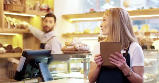 Close-up van de jonge blanke mooie vrouwelijke verkoper in schort staan in de bakkerij, tikken en typen op tablet apparaat, terwijl mannelijke collega werkt op de achtergrond. Bakkerij concept — Stockvideo