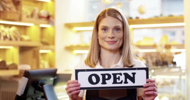 Sluit maar af. Vrolijke blanke mooie jonge vrouw staan in eigen bakkerij en houden Open teken in de handen, kijken naar de camera en glimlachen. De bakkerij gaat weer open. Zakenvrouw opent winkel — Stockvideo