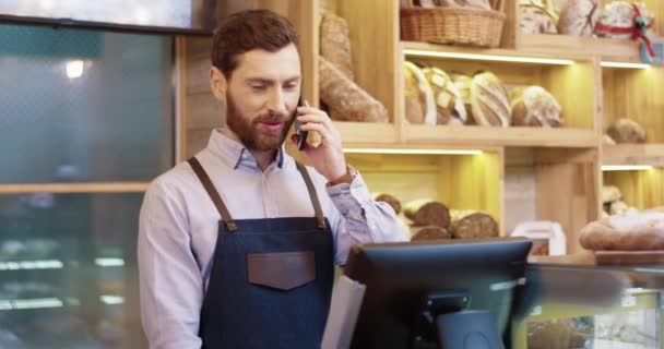 カウシャ人の髭を生やしたハンサムな男のパン屋さんがカウンターでパン屋に立って携帯電話で話していた。男性労働者は顧客と携帯電話で話す。パン屋のコンセプト — ストック動画