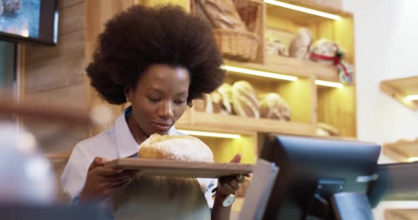 Sluiten van vrolijke mooie jonge Afro-Amerikaanse vrouw verkoper in eigen bakkerij in schort staan en houden lade met vers gebakken brood in handen, ruiken bakken en glimlachen naar de camera — Stockvideo