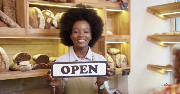 Primer plano retrato de joven alegre afroamericana bonita vendedora en delantal sosteniendo Cartel abierto en las manos de pie en una pequeña panadería de buen humor al reabrir, mirando a la cámara y sonriendo — Vídeos de Stock