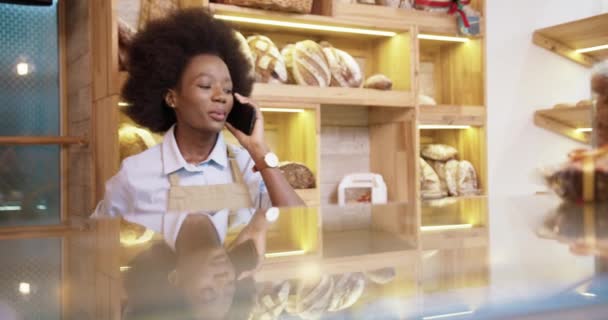 Primer plano retrato de alegre joven afroamericana bonita vendedora de pie en una pequeña panadería de buen humor llamando y hablando en el teléfono inteligente en el lugar de trabajo. Concepto de trabajador de panadería — Vídeos de Stock