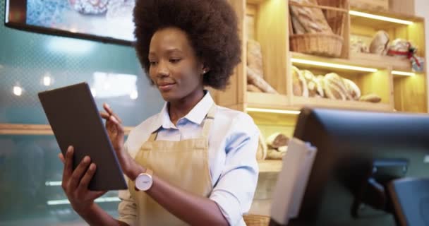 Primer plano retrato de alegre joven afroamericana hermosa trabajadora vendedora de pie en pequeña panadería de buen humor mecanografía y desplazamiento en la tableta en el lugar de trabajo. Concepto de trabajador de panadería — Vídeos de Stock