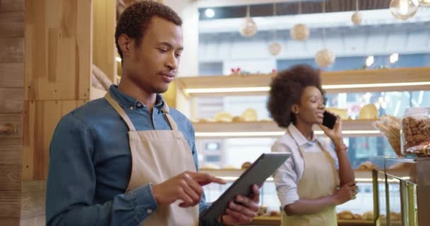 Vista lateral. Close up de belo jovem afro-americano trabalhador homem na padaria digitação e navegação em tablet enquanto colega de trabalho do sexo feminino falando no celular no trabalho. Conceito de negócio. Empresários a trabalhar — Vídeo de Stock