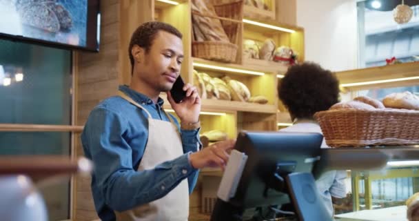 Großaufnahme eines fröhlichen jungen, gut aussehenden afroamerikanischen Arbeiters in Schürze, der in der Backstube mit dem Handy telefoniert, während eine Kollegin arbeitet und Brot in die Regale stellt. Unternehmerkonzept — Stockvideo