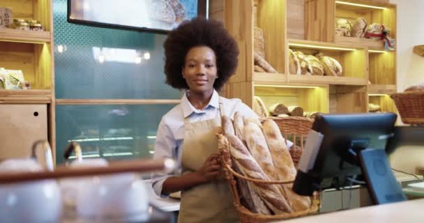 Feliz hermosa joven empresaria afroamericana en delantal se encuentra en su propia pequeña panadería con pan fresco baguettes en las manos y sonriendo a la cámara. Concepto empresarial — Vídeos de Stock