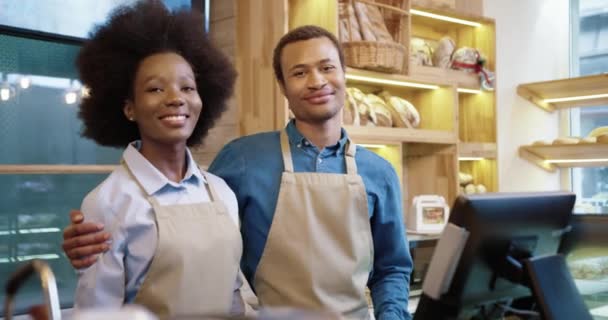 Портрет счастливой афроамериканской пары, ведущей семейный бизнес. Мужчина и женщина-пекари в фартуках стоят в пекарне и улыбаются, обнимаясь перед камерой. Маленькая собственная пекарня. Концепция бизнеса — стоковое видео