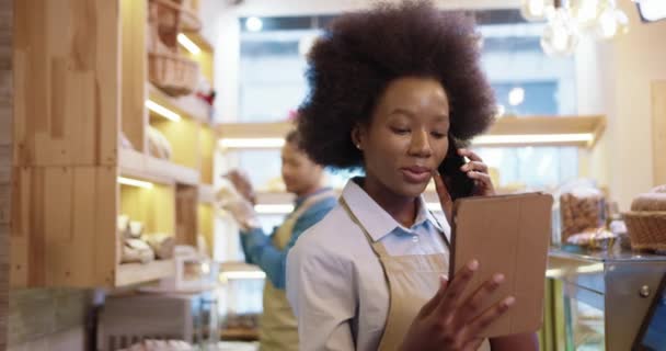 De cerca. Afroamericana hermosa mujer feliz en delantal llamando y hablando por teléfono móvil usando tableta mientras está de pie en la panadería. Su compañera de trabajo trabajando en el fondo. Concepto de panadería — Vídeos de Stock