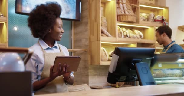 Gros plan de joyeux souriant afro-américaine belle femme dans le tablier en utilisant la tablette tapant et tapant tout en se tenant dans la boulangerie parler à son collègue homme. Concept de boulangerie — Video