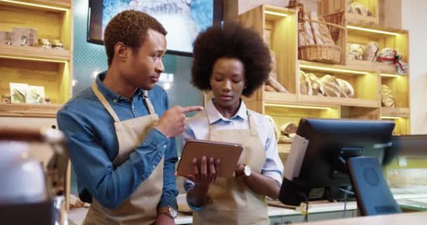 Ritratto di felice sorridente giovane coppia afroamericana uomo e donna in grembiuli utilizzando tablet toccando e digitando mentre in piedi nel proprio negozio di panetteria parlando al coperto. Concetto di panetteria — Video Stock