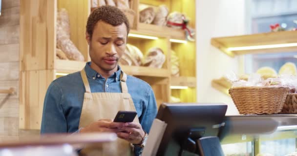 Detailní záběr mladého afroamerického pohledného mužského pracovníka v zástěře stojí v pekárně, píše a píše na smartphone. Muž prodejce procházení on-line na mobilu. Obchodní koncept. Maloobchod — Stock video