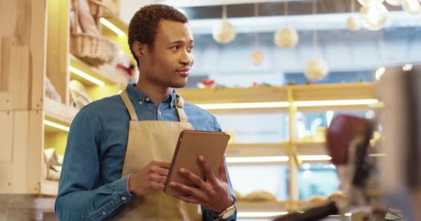 アフリカ系アメリカ人の幸せな若いハンサムな男性労働者のクローズアップエプロンでパン屋さんに立って、テキストメッセージやタブレットを入力します。男性の売り手はデバイス上でオンラインで閲覧。ビジネスコンセプト。小売業 — ストック動画