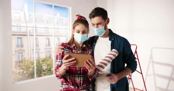 Retrato de la joven pareja casada caucásica feliz hombre y mujer en máscaras de pie en el apartamento escribiendo en el dispositivo de la tableta discutiendo el diseño de la nueva habitación. Concepto de reparación del hogar. decoración y renovación — Vídeos de Stock