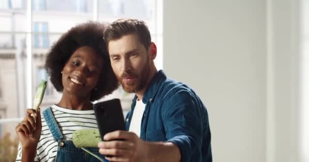 Πορτρέτο των ευτυχισμένων μικτών αγώνων νεαρό παντρεμένο ζευγάρι οικογένειας άνδρας και γυναίκα χαμογελώντας σε νέο διαμέρισμα λαμβάνοντας φωτογραφίες selfie στο smartphone ποζάρουν με βούρτσες στο δωμάτιο. Αρχική ιδέα επισκευής — Αρχείο Βίντεο