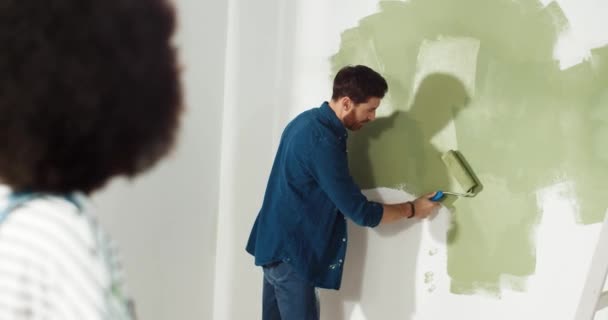 Parte trasera de la mujer afroamericana de pie en casa y mostrando a su marido caucásico cómo reparar y redecorar la habitación. Hombre pintando pared con rodillo de cepillo y sonriendo. Renovación del concepto plano — Vídeos de Stock