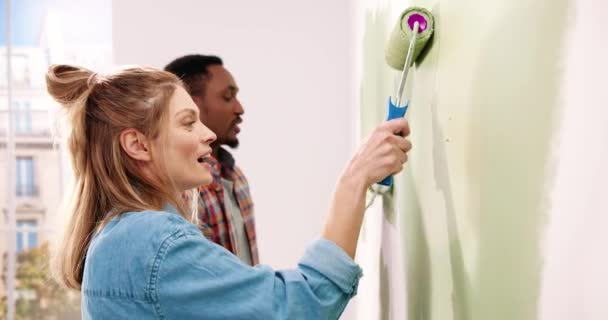 Primer plano retrato de la feliz joven pareja de razas mixtas de la familia juntos en el nuevo apartamento renovando la pared de la pintura de la habitación con rodillo de cepillo en las manos. renovar el concepto de hogar. Vista lateral Decoración y reparación — Vídeos de Stock