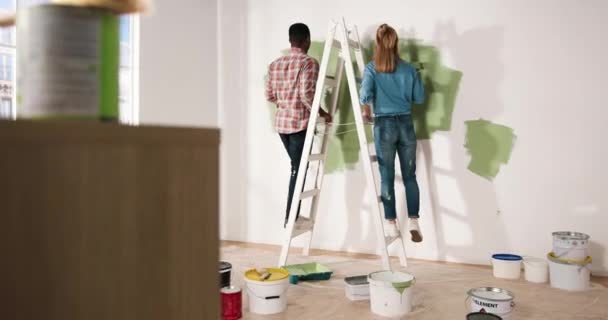 Vista posterior de la pareja multi-étnica hombre afroamericano y amigos de la mujer caucásica de pie en la escalera de paso de reparación y redecoración de la habitación. Paredes de pintura familiar con rodillo de pincel en nuevo color. — Vídeos de Stock