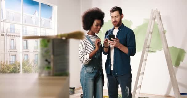 Joyful young married couple Caucasian husband and African American wife standing in room typing on smartphone choosing design of new home. Excited man and woman choosing decor for home renovation — Stock Video