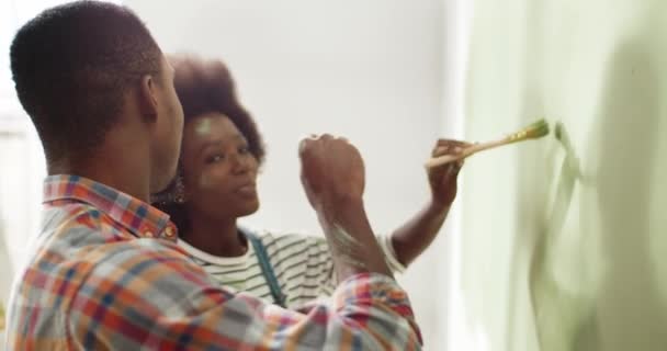 Κοντινό πλάνο του χαριτωμένο παντρεμένος ευτυχισμένος νεαρός Αφρικής ζευγάρι των ΗΠΑ ζωγραφική τοίχους στο διαμέρισμα σε νέο χρώμα με πινέλο και τη διασκέδαση. Σύζυγος και σύζυγος γελούν και ζωγραφίζουν ο ένας πάνω στον άλλον — Αρχείο Βίντεο
