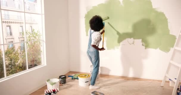 Joven mujer afroamericana bailando y pintando pared con pincel de rodillo mientras renueva apartamento. Parte trasera de la hembra que se divierte redecorando el hogar, renovando y mejorando el concepto de reparación y decoración — Vídeos de Stock