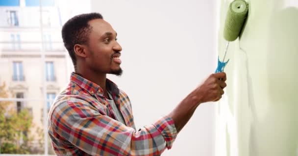 Zijaanzicht. Afro-Amerikaanse klusjesman schilderen muur met roller borstel tijdens woningrenovatie. Appartement renovatie en woningbouw tijdens de renovatie en verbetering. Reparatie en decoratie — Stockvideo