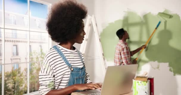 Portrait of happy pretty African American woman typing on laptop in room during home repair speaking with husband who is painting walls in olive color with roller brush. House renovation and redesign — Stock Video