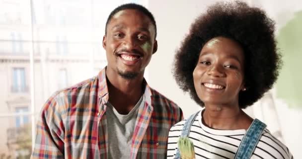 Joyful young cute African American couple looking at camera and smiling standing indoor in room with paint brush during home repair process. wife and husband doing renovation in house Close up concept — Stock Video