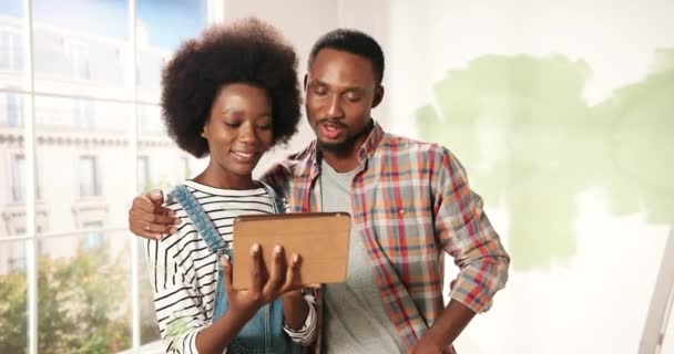 Alegre lindo afroamericano joven esposa y marido pareja golpeteo y navegación en el dispositivo de la tableta elegir ideas de decoración para el nuevo apartamento discutir el rediseño de la casa. Reparación y mejoras en el hogar — Vídeo de stock