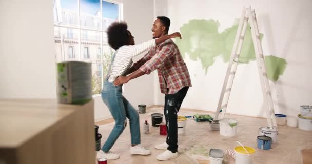 Jovem alegre casal afro-americano homem e mulher abraçando e abraçando de bom humor sorrindo no apartamento durante as obras de reparação em casa. Esposa e marido renovando casa. Conceito de remodelação — Vídeo de Stock