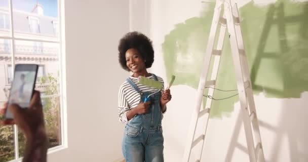 Afro-americano encantador marido tirar fotos de bela esposa feliz no quarto fazendo renovação em casa posando com pincel nas mãos. Renovação e conceito de reparação em casa. Fotografar no celular — Vídeo de Stock