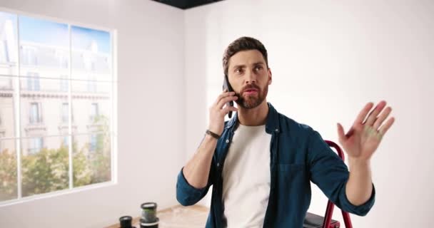 Handsome young joyful Caucasian handyman speaking on mobile phone with client in new apartment. Happy male chatting on smartphone during home repair and renovation. Redesigning room concept — Stock Video
