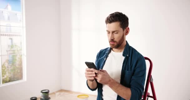 Portrait of Caucasian happy young bearded man sitting on ladder in empty apartment during repair works texting and tapping on cellphone. Renovation and improvement concept. Redesigning house concept — Stock Video