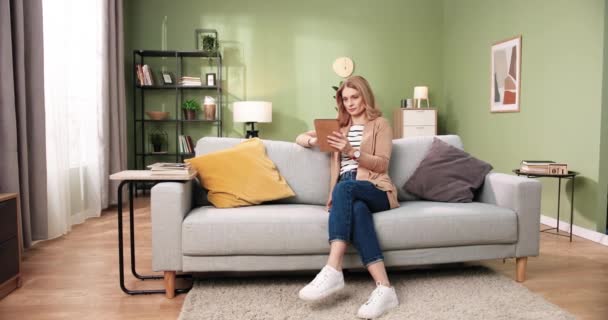 Modern, rahat bir dairede kanepede oturan güzel beyaz kadın tablet cihazıyla internette geziniyor. Oturma odasında dinlenen neşeli, güzel bir kadın. Boş zaman konsepti — Stok video
