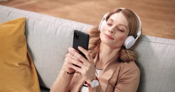 Joven mujer caucásica alegre que se relaja en casa sentada en el sofá usando el teléfono inteligente que navega por Internet y escuchando la canción favorita de la música en los auriculares en el teléfono celular moderno en casa. Concepto de primer plano — Vídeos de Stock