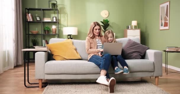 Joven madre e hija caucásicas usando el portátil para la educación sentado en el sofá en casa durante la cuarentena. Hermosa mujer y niño mirando la pantalla de la computadora con sonrisas felices navegando en línea — Vídeos de Stock