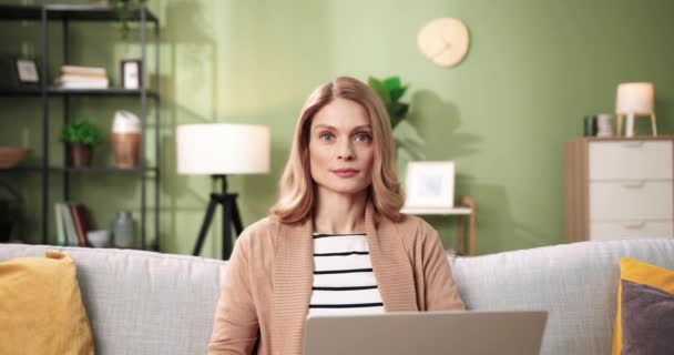 Retrato de alegre hermosa mujer caucásica de buen humor se sienta en el sofá en acogedora habitación moderna con ordenador portátil, mirando a la cámara y sonriendo. Trabajando lejos de casa. Ocio en interiores. Concepto casero — Vídeo de stock