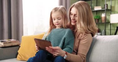 Neşeli, güler yüzlü beyaz bir annenin küçük tatlı kızıyla vakit geçirmesi tablet cihazında yazı yazmayı öğretmesi, internette kanepeye oturması portresi. Annelik kavramı. Mutlu çocukluk