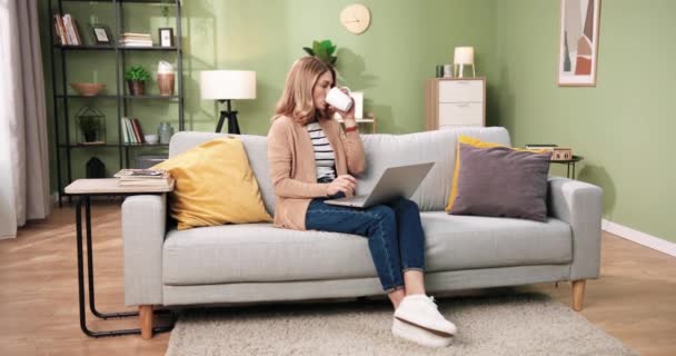 白种人漂亮的年轻女性坐在舒适的客厅沙发上，在公寓打字，在笔记本电脑上工作，一边喝热咖啡。离家的距离工作。休闲的概念。现代公寓 — 图库视频影像