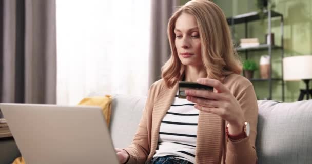 Vacker vit ung kvinna med nöjda ansikte skriva och bläddra på laptop, vilket gör inköp på internet håller kreditkort i handen och ler i rummet. e-handel — Stockvideo