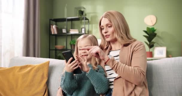 Retrato de la alegre madre cariñosa de la familia caucásica y la pequeña hija linda pasar tiempo juntos sentados en el sofá escribiendo y navegando en línea en el teléfono inteligente Concepto de maternidad Las relaciones de mamá y niña — Vídeos de Stock