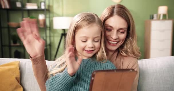 Närbild. Glada söt trevlig mamma sitter med liten förskola dotter på soffan i mysiga rum och talar på nätet videosamtal på surfplatta, viftande händer och leende i positiv stämning. Barndom — Stockvideo