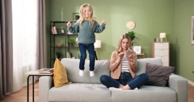 Beyaz tenli güzel bir kadın kanepeye oturmuş akıllı telefonuyla mesajlaşıyor sosyal medyayı kullanıyor ve küçük tatlı kızı da evdeki kanepeye atlıyor. Çocukluk anneliği kavramı