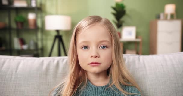 Κοντινό πορτρέτο του καυκάσιου μικρού αξιολάτρευτου κοριτσιού που κάθεται στον καναπέ του δωματίου, κοιτάζοντας την κάμερα και χαμογελώντας με θετική διάθεση. Ευτυχισμένη παιδική ηλικία. Μικρό χαριτωμένο παιδί προσχολικής ηλικίας στο σπίτι μόνο — Αρχείο Βίντεο