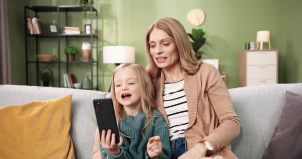 Portrait de joyeux jolie mère attentionnée caucasienne assise avec une petite fille maternelle sur un canapé dans une pièce confortable et bavardant sur un appel vidéo en ligne sur un téléphone portable, agitant les mains et souriant. Concept de maison — Video